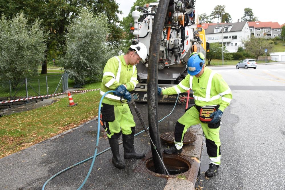 Two workers in high-visibility gear clean a sewer using a vacuum truck and specialized tools.