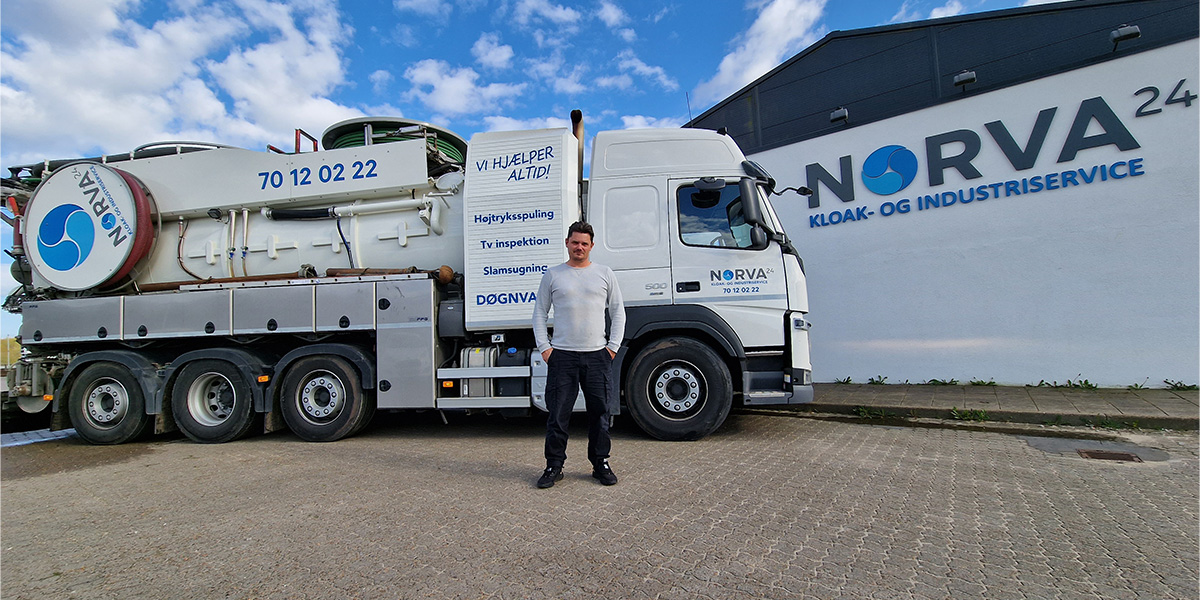 Man stands by a Norva24 truck with branding, parked outside the company building on a sunny day.