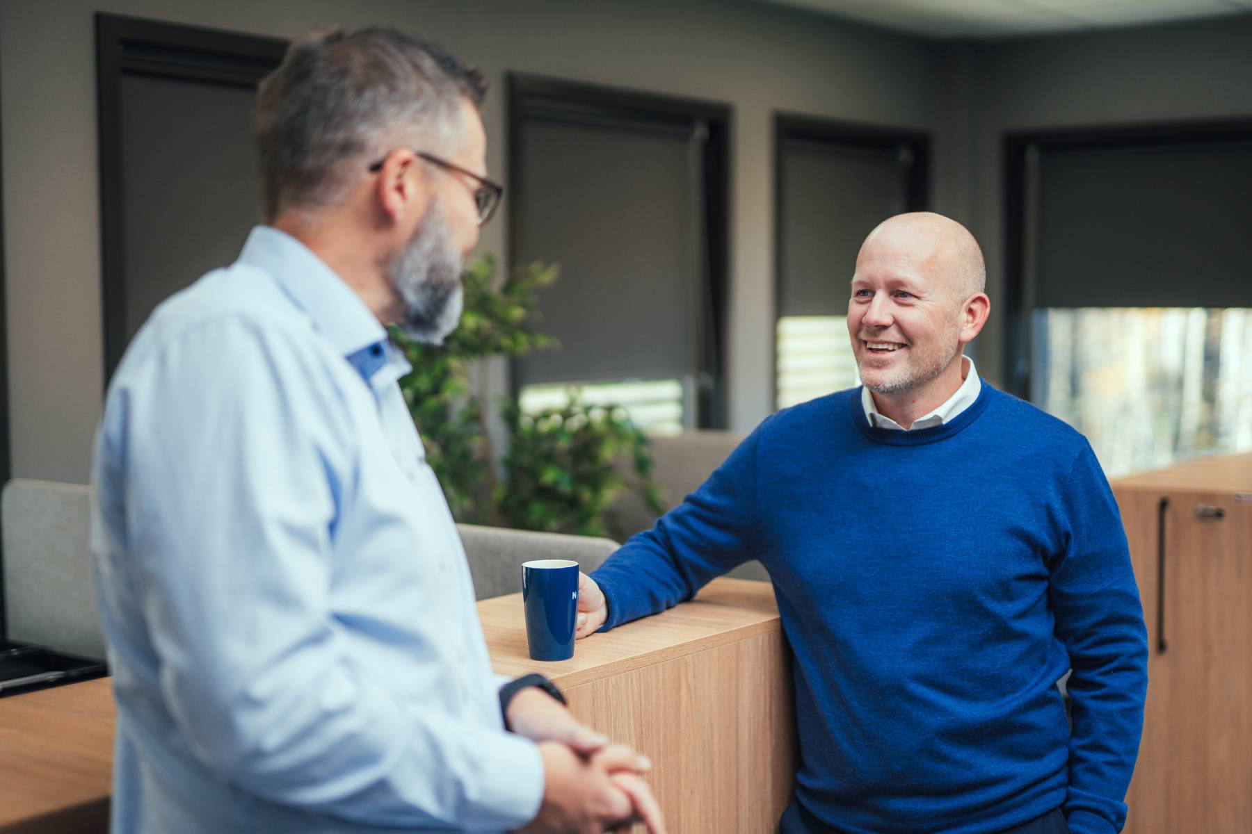 Two men in an office having a conversation, one leaning on a wooden desk with a coffee cup.