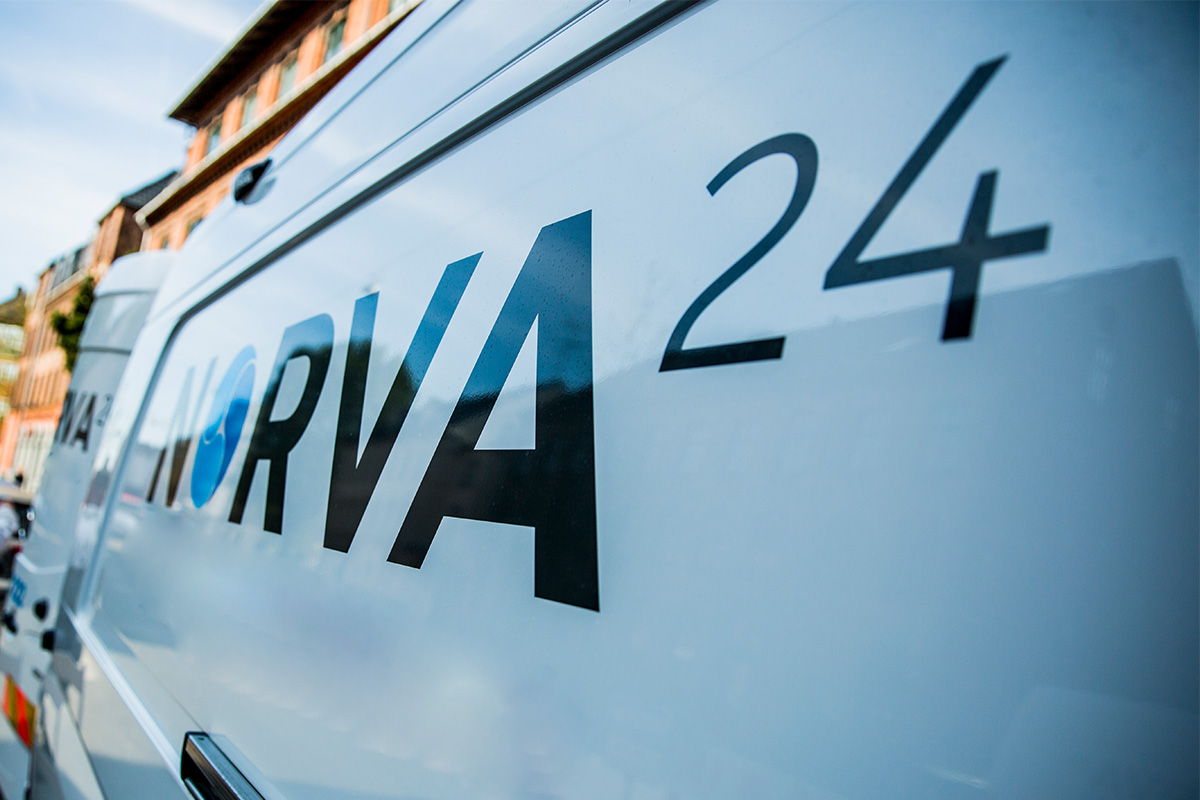 Close-up of the Norva24 logo on the side of a white service van with a blurred urban background.